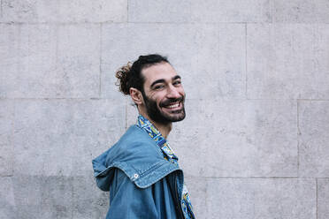 Smiling young man with denim jacket standing in front of wall - XLGF01291