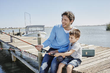 Cheerful father fishing with son while sitting on pier - RORF02670