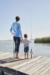Father and son with fishing rods holding hands while standing on pier - RORF02671