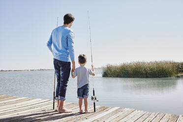 Father and son holding hands while standing with fishing rods on jetty - RORF02672