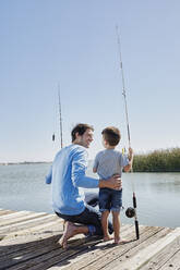 Smiling father looking at son standing with fishing rod on pier - RORF02673