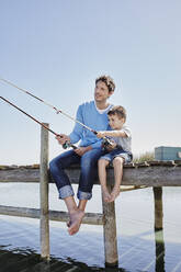 Mature man and boy sitting with fishing rods on pier - RORF02677