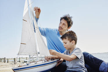 Vater und Sohn bereiten gemeinsam ein Spielzeug-Segelboot vor, während sie am Pier sitzen - RORF02680