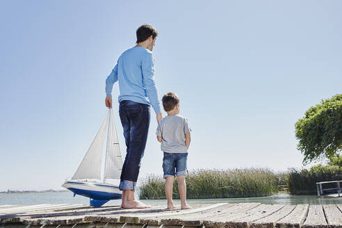 Älterer Mann mit Spielzeugsegelboot, der die Hand eines Jungen hält, während er gegen den Himmel steht - RORF02682