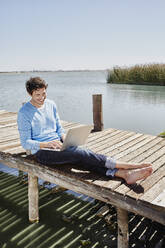Smiling man using laptop while sitting on pier during sunny day - RORF02684