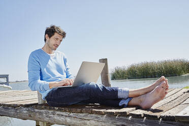 Mann, der einen Laptop benutzt, während er an einem sonnigen Tag auf einem Steg sitzt - RORF02685