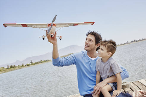 Vater hält Flugzeugspielzeug, während er mit seinem Sohn am See sitzt - RORF02697