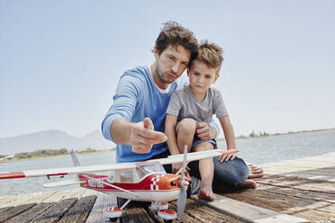 Vater spricht mit seinem Sohn über Flugzeugspielzeug, während er auf dem Pier sitzt - RORF02698