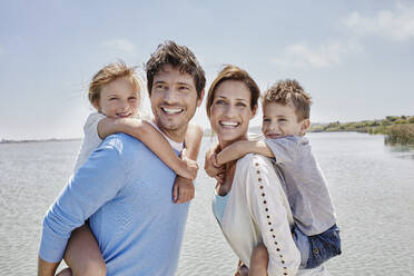 Lächelnde Eltern, die ihre Kinder an einem sonnigen Tag am See huckepack nehmen - RORF02704