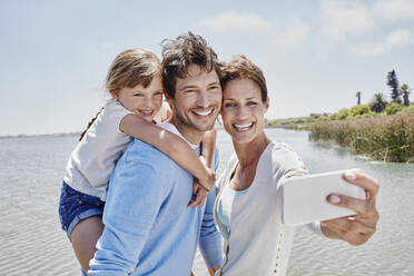 Smiling woman taking selfie with family by lake on sunny day - RORF02706