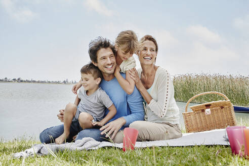 Glückliche Familie sitzt in einer Reihe auf einer Decke am See - RORF02711
