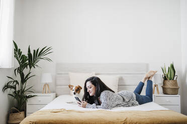 Lächelnde Frau, die ein Smartphone mit einem Jack Russell Terrier benutzt, während sie zu Hause auf dem Bett liegt - EBBF02607
