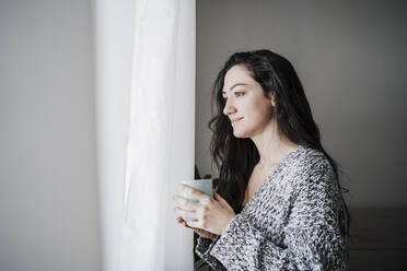 Thoughtful smiling woman holding coffee cup while looking through window at home - EBBF02620