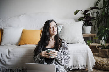 Lächelnde Frau mit Laptop, die eine Kaffeetasse hält und auf dem Boden vor dem Sofa im Wohnzimmer sitzt - EBBF02622