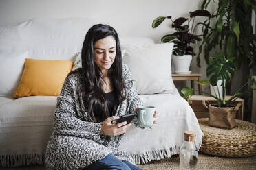 Smiling woman holding coffee cup while using mobile phone against sofa in living room at home - EBBF02623