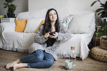 Smiling woman looking away while using smart phone against sofa in living room - EBBF02625