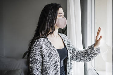 Woman with protective face mask looking through window while standing in living room at home during COVID-19 - EBBF02631