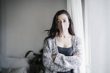 Woman wearing protective face mask standing by window at home - EBBF02632