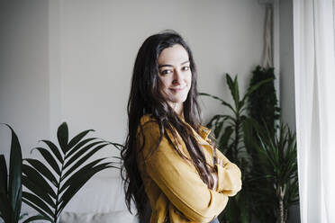 Smiling woman in yellow coat standing with arms crossed in living room at home - EBBF02635