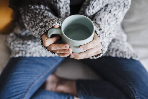 Frau hält Becher mit Wasser, während sie zu Hause auf dem Sofa sitzt - EBBF02645