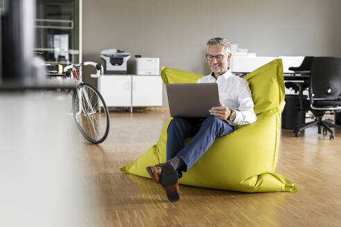 Smiling businessman using laptop while sitting on beanbag in office - PESF02638