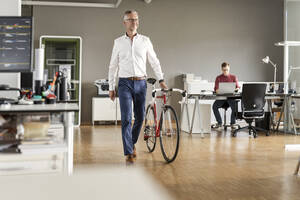 Älterer Geschäftsmann mit Fahrrad in einem Großraumbüro - PESF02639