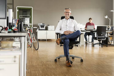 Lächelnder Geschäftsmann mit gekreuzten Beinen auf einem Stuhl im Büro sitzend - PESF02641