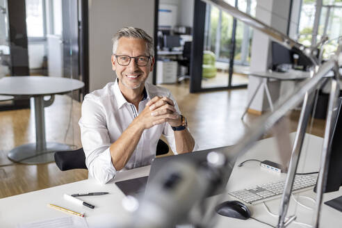 Smiling businessman with hands clasped sitting at desk in office - PESF02646