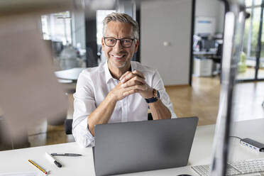 Lächelnder männlicher Unternehmer mit Laptop, der in einem Großraumbüro am Schreibtisch sitzt - PESF02651