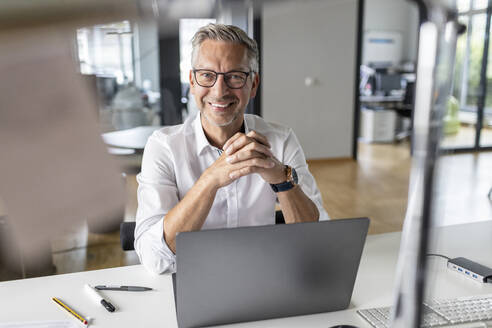 Smiling male entrepreneur with laptop sitting at desk in open plan office - PESF02651