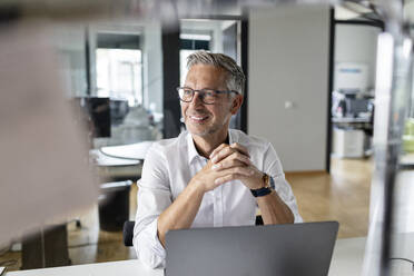 Smiling businessman with hands clasped looking away while sitting at desk in office - PESF02652