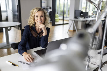 Beautiful smiling businesswoman with hand on chin sitting at desk in office - PESF02653