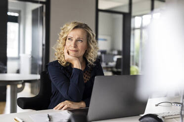Thoughtful female business professional with head in hand looking away while sitting at desk in office - PESF02656