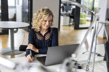 Lächelnde Geschäftsfrau, die einen Stift hält und auf einen Laptop im Büro schaut - PESF02659