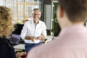 Smiling male business professional holding digital tablet while discussing with colleagues in office - PESF02671