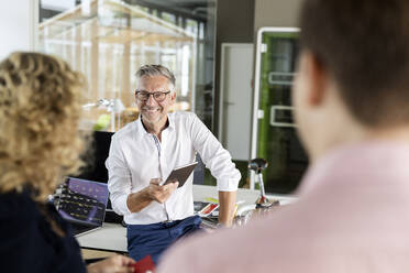 Smiling businessman with digital tablet discussing with colleagues at desk in office - PESF02672