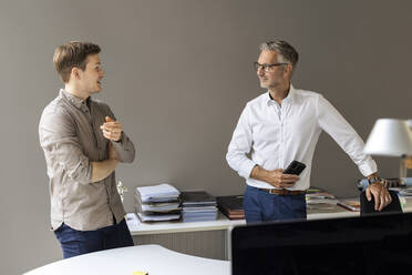 Young businessman talking with mature colleague while standing against wall in office - PESF02678