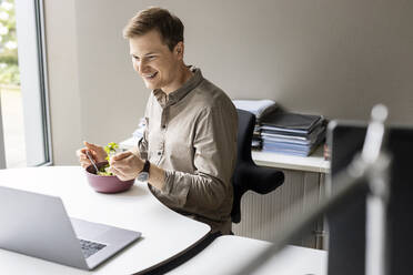 Lächelnder junger Geschäftsmann beim Mittagessen mit Blick auf den Laptop im Büro - PESF02680