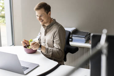 Smiling young businessman having lunch while looking at laptop in office - PESF02680