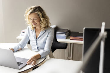 Smiling businesswoman with laptop sitting at desk in office - PESF02681