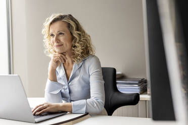 Smiling businesswoman with hand on chin looking away while sitting in office - PESF02682