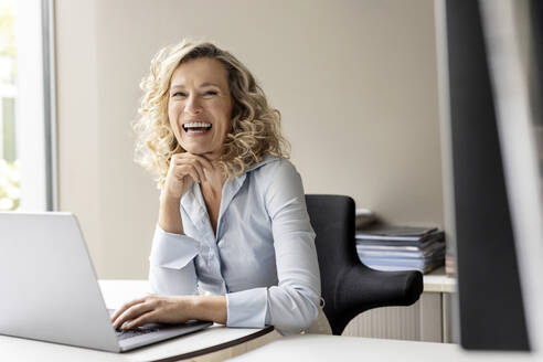 Laughing female business professional with laptop sitting in office - PESF02685
