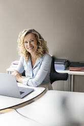 Smiling female business professional with laptop sitting at desk in office - PESF02686