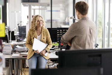 Smiling businesswoman with laptop discussing with young colleague while leaning at desk in office - PESF02698