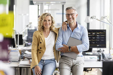 Smiling businessman with digital tablet leaning on desk by businesswoman in office - PESF02705