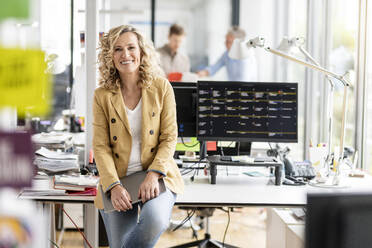 Lächelnde Geschäftsfrau mit digitalem Tablet, die sich an einen Computer am Schreibtisch im Büro lehnt - PESF02709