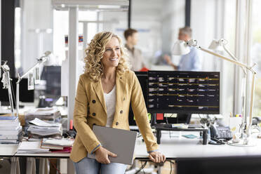 Lächelnde Geschäftsfrau, die wegschaut, während sie ein digitales Tablet am Schreibtisch im Büro hält - PESF02710