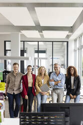 Smiling businesswoman with digital tablet standing amidst male and female colleagues against desk in office - PESF02713