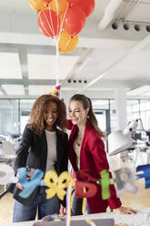 Lächelnde Geschäftsfrau hält ein Bündel Luftballons, während sie mit einer Kollegin im Büro Geburtstag feiert - PESF02718