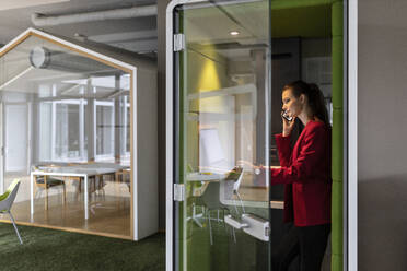 Smiling female entrepreneur using laptop while talking on mobile phone in soundproof cabin at office - PESF02730